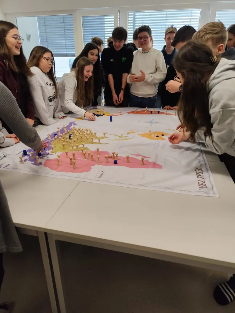 Eine Gruppe von Schülern ist um eine große Karte auf einem Tisch versammelt, möglicherweise an einer Klassenaktivität beteiligt. Sie tragen Freizeitkleidung und einige lächeln.