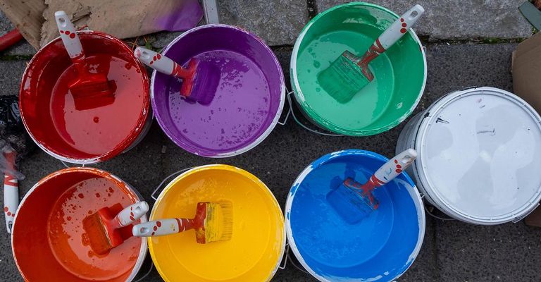 Mehrere Eimer mit verschiedenen Farben von Farbe stehen auf dem Boden, wobei in jedem ein Pinsel steckt.