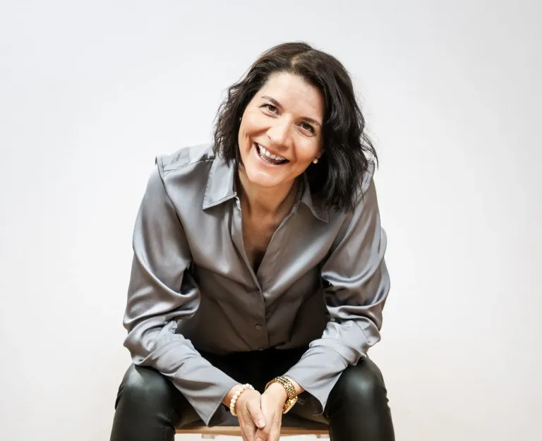 Eine lächelnde Frau mit kurzen schwarzen Haaren, die eine graue Bluse und schwarze Hose trägt, sitzt auf einem Stuhl und posiert möglicherweise für ein Foto.