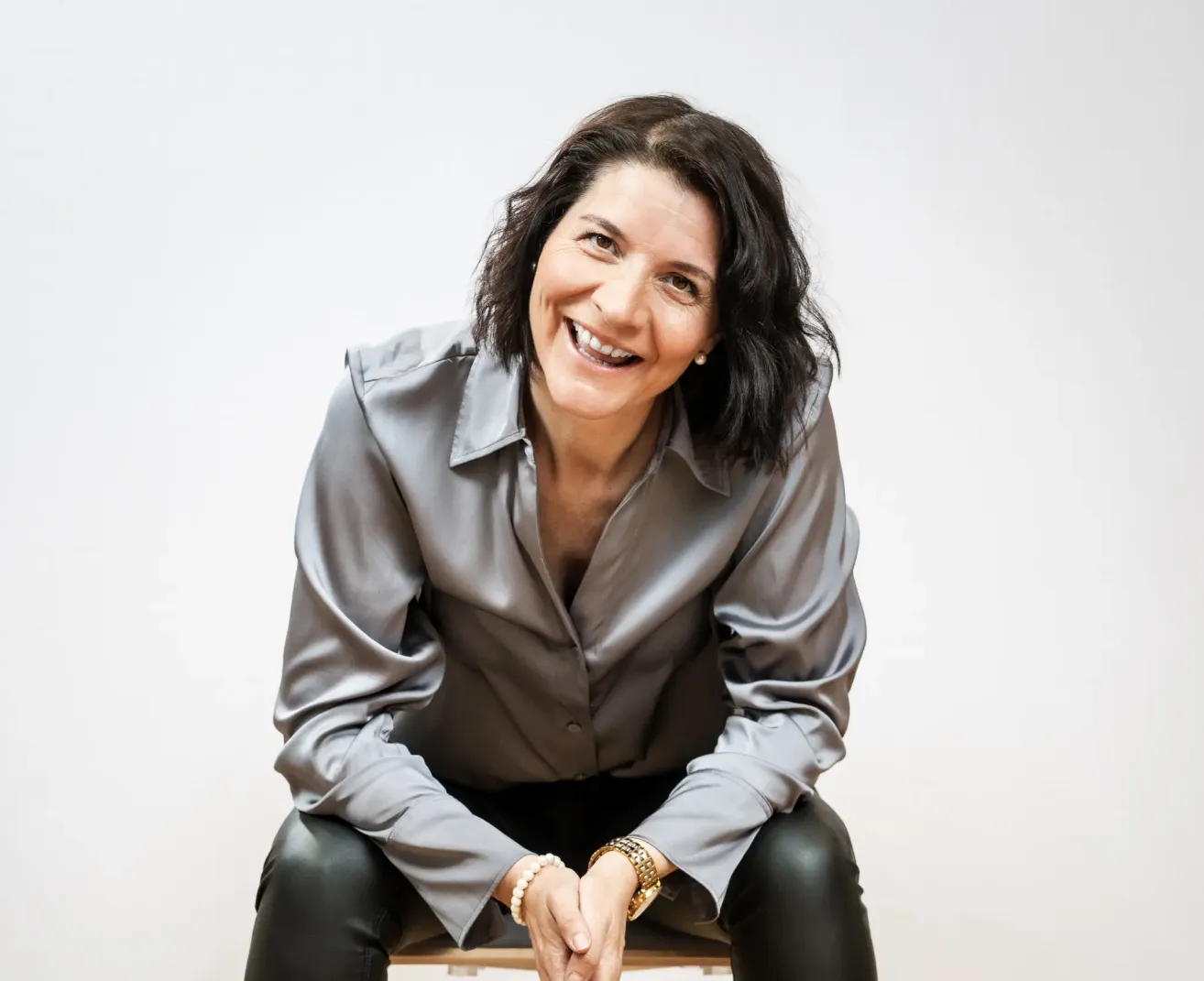 Eine lächelnde Frau mit kurzen schwarzen Haaren, die eine graue Bluse und schwarze Hose trägt, sitzt auf einem Stuhl und posiert möglicherweise für ein Foto.