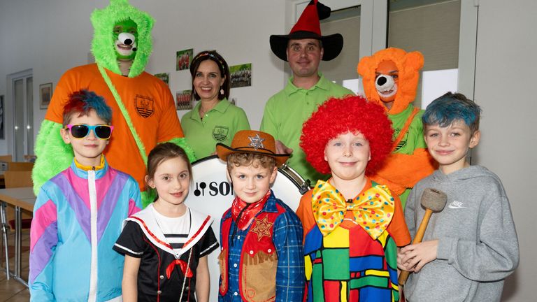 Eine Gruppe von Kindern und Erwachsenen posiert für ein Foto, einige in Kostümen. Ein Mädchen in einer Matrosenuniform steht neben einem Jungen in einem Cowboyhut. Dahinter lächeln ein Mann in einem grünen Poloshirt und eine Frau in einem grünen Poloshirt. Ein Junge in einem Clownskostüm und ein Mädchen in einem Bärenkostüm sind ebenfalls anwesend.