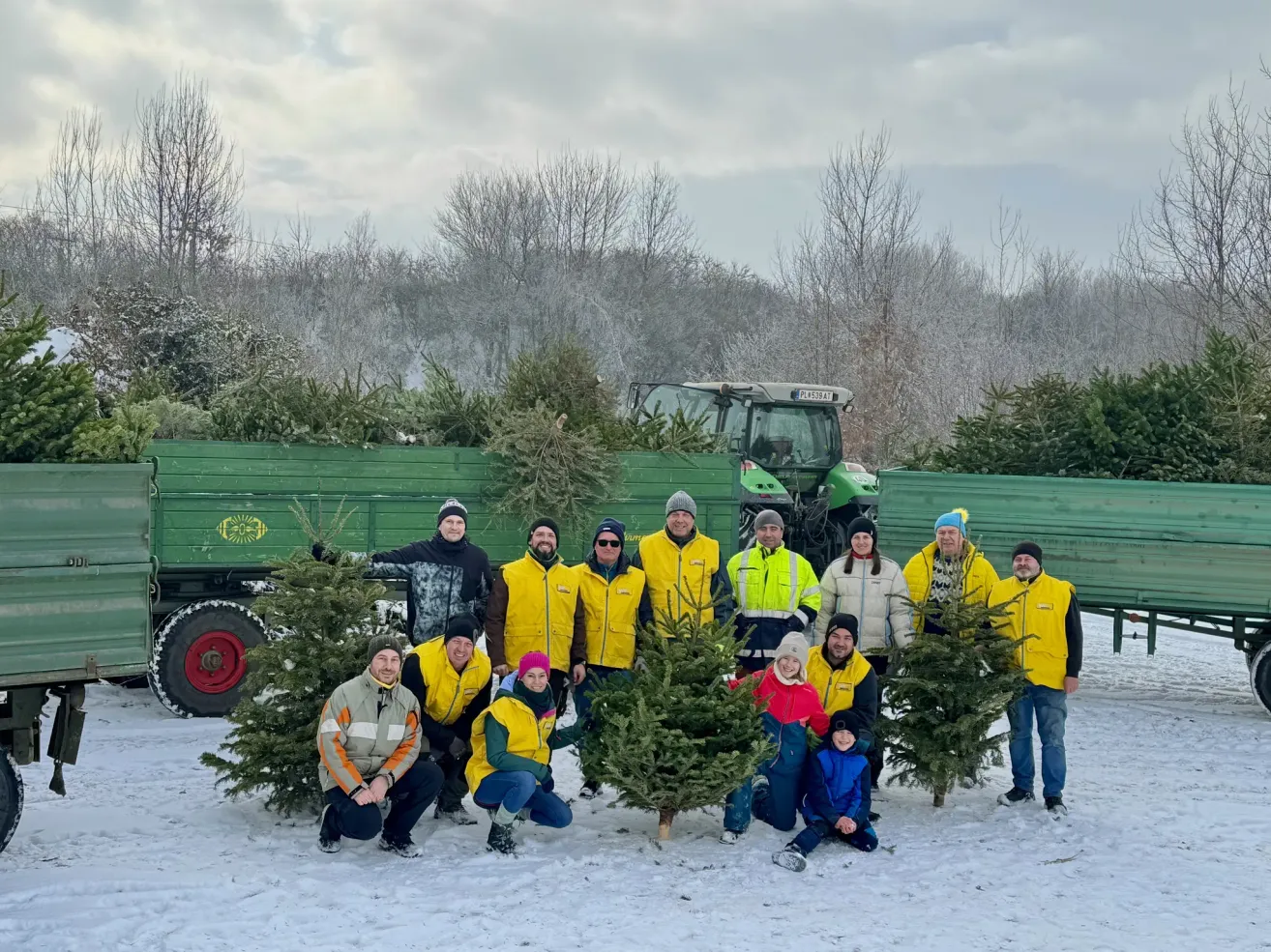 Eine Gruppe von Menschen in Winterjacken posiert für ein Foto im Schnee mit Tannenbäumen. Dahinter sind zwei grüne Lastwagen mit Traktor geparkt.