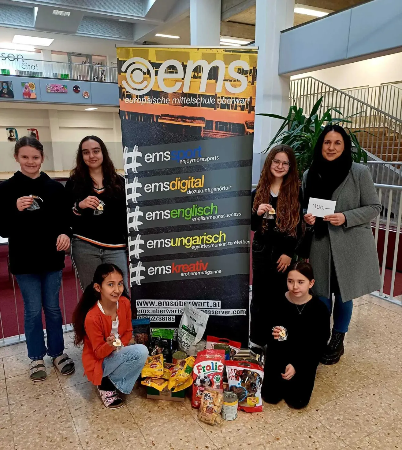 Fünf Mädchen stehen vor einem Schild und lächeln. Sie halten Medaillen und posieren neben einem Banner. Dahinter steht eine Treppe und links eine Brüstung. Auf dem Boden liegen Essen und Snacks.