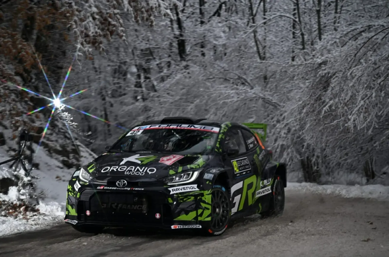 Ein Rallye-Auto mit grüner und schwarzer Lackierung fährt auf einer verschneiten Straße. Es hat einen Sponsorenaufkleber mit der Aufschrift 'Organiq Wood'.