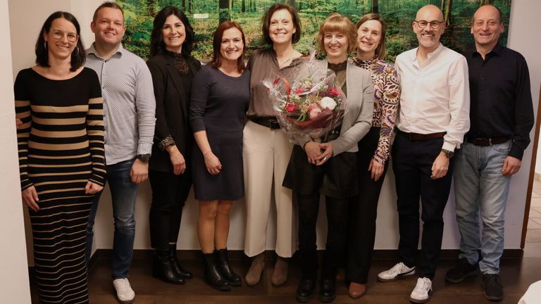 Eine Gruppe von sieben Menschen posiert für ein Foto vor einer Wand mit einem Waldgemälde. Sie lächeln. Eine Frau in der Mitte hält einen Blumenstrauß.