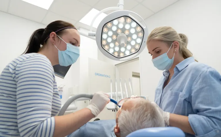 Zwei Zahnärzte in Masken untersuchen die Zähne eines älteren Patienten. Einer benutzt einen Zahnspiegel, während der andere ein Dentalwerkzeug hält. Der Schauplatz ist eine Zahnarztpraxis.