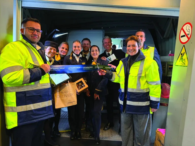 Eine Gruppe von Menschen, einschließlich Mitarbeiter, hält ein Band mit der Aufschrift 'Lufthansa Group Taste & More'. Sie stehen in einem Fahrzeug, möglicherweise einem Lieferwagen, mit Tüten voller Lebensmittel.