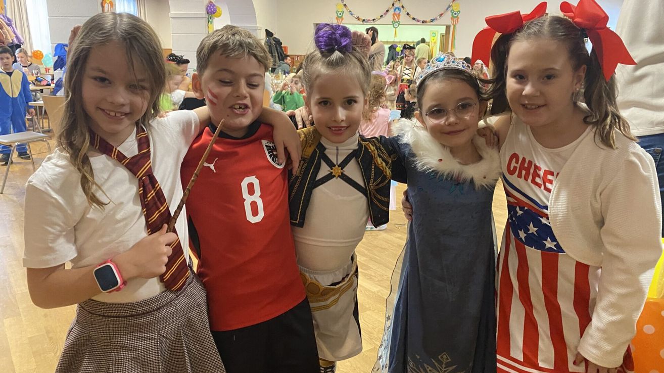 Eine Gruppe von Kindern in Kostümen, darunter ein Junge in einem roten Trikot mit der Nummer 8, ein Mädchen in einem weißen Kostüm und ein weiteres Mädchen in einem blauen Kleid.