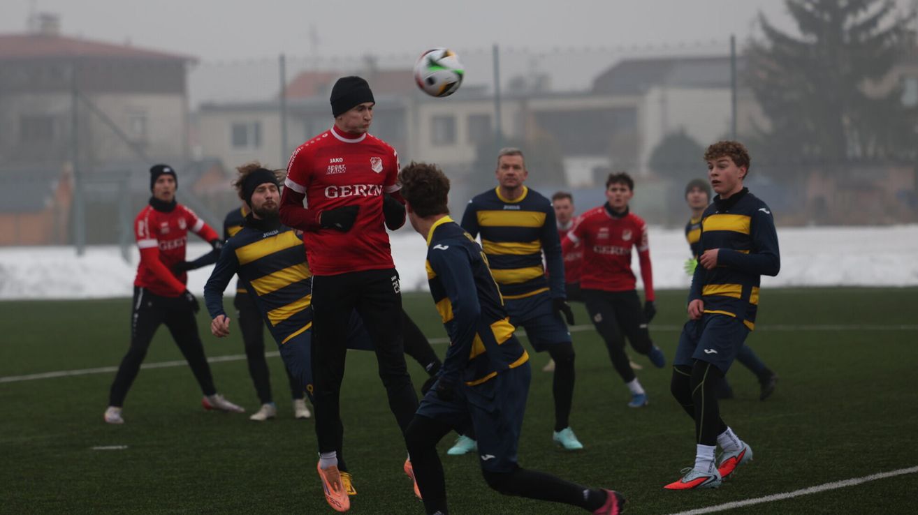 Fußballspieler konkurrieren auf einem nebligen Feld. Ein Spieler in Rot springt, um den Ball zu köpfen. Andere schauen zu und sind bereit zu reagieren. Sie tragen passende Uniformen, einige in Blau und Gelb.