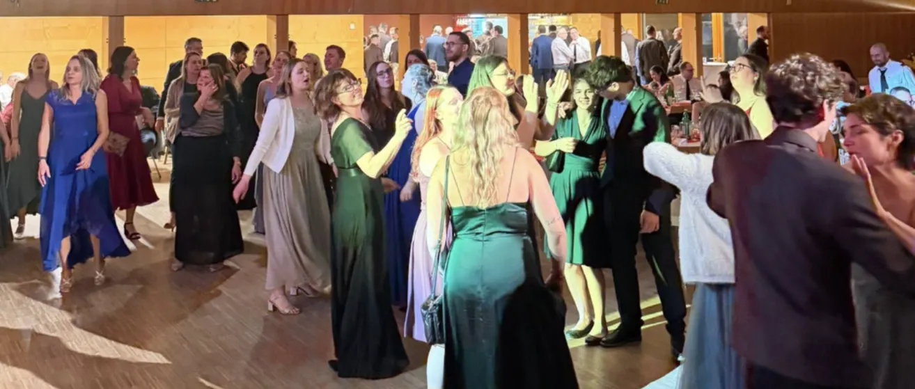 Eine Gruppe von Menschen in formeller Kleidung steht und tanzt in einem Saal. Eine Frau mit Brille klatscht in die Hände.