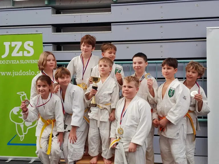 Eine Gruppe junger Kampfkünstler in weißen Uniformen und Medaillen posiert für ein Foto in einer Sporthalle. Ein Junge hält einen Pokal.