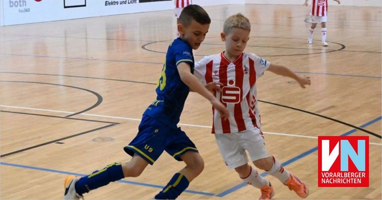 Zwei junge Jungen in Fußballtrikots spielen auf einem Hallenplatz. Der Junge in Blau hat die Nummer 3 auf seinem Trikot, während der Junge in Rot und Weiß die Nummer 5 hat. Sie schauen beide konzentriert, während sie spielen.