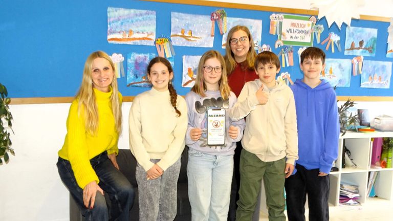 Eine Gruppe von Schülern und einer Lehrerin stehen vor einer Tafel mit Kunstwerken. Die Schüler halten eine Plakette, und die Lehrerin lächelt. Sie tragen alle Freizeitkleidung.