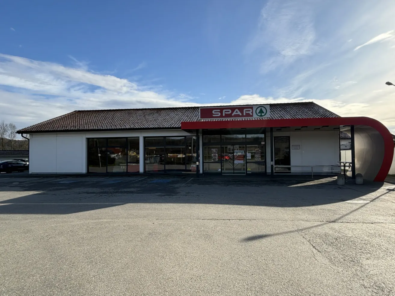 Das Äußere eines Spar-Geschäfts. Es hat ein braunes Dach, ein rotes Vordach und große Glasfenster. Vor dem Gebäude befinden sich einige Bänke und ein Tisch.