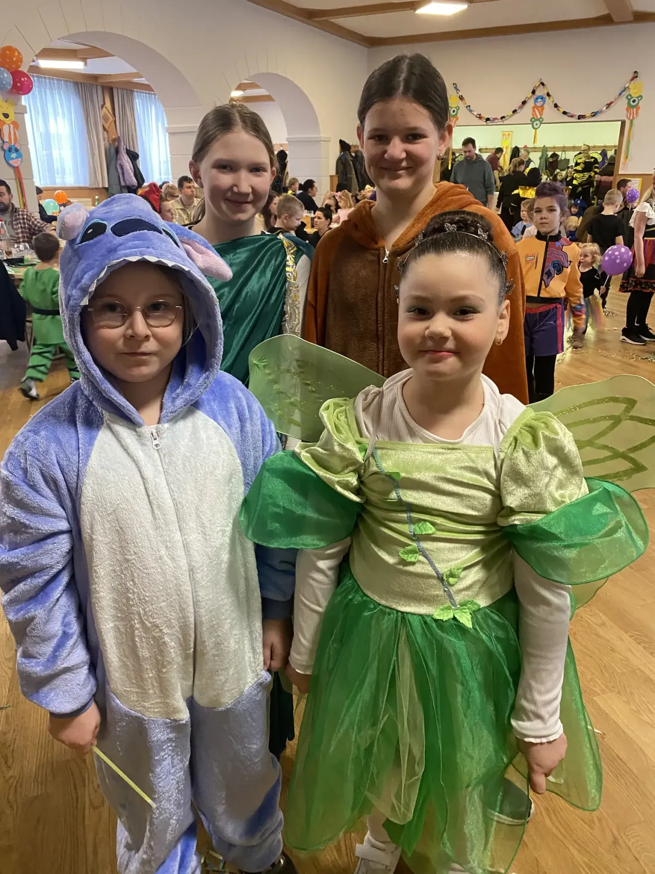 Vier Kinder in Kostümen, darunter ein blauer Stitch, ein grüner Feenflügel, eine braune Kapuze und eine grüne Toga, posieren für ein Foto in einem Raum mit Holzböden.