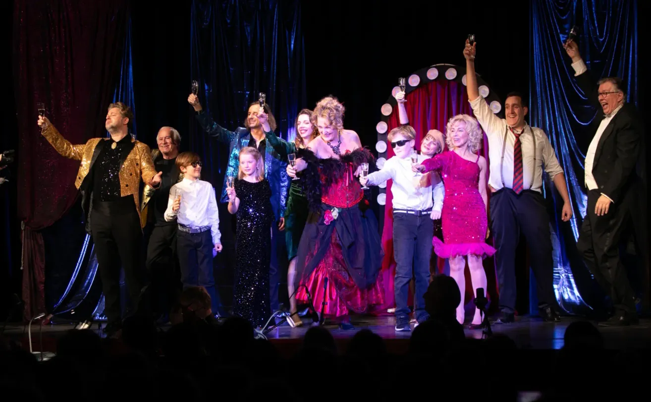 Eine Gruppe von Darstellern feiert auf einer Bühne mit Champagnergläsern, umgeben von einem blauen Vorhang und einem roten Hintergrund.
