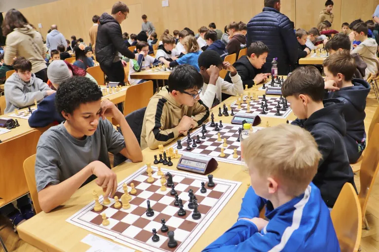 Eine Gruppe von Kindern spielt Schach in einem Raum mit Holzwänden. Es gibt viele Schachbretter auf dem Tisch mit Spielfiguren. Die Kinder schauen auf die Schachbretter.