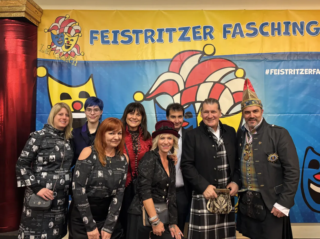 Eine Gruppe von Erwachsenen in Kostümen steht vor einem Banner mit dem Text 'Feistritzer Fasching'. Sie scheinen für ein Foto zu posieren.
