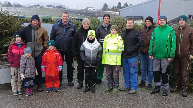 Eine Gruppe von Menschen steht in einer Reihe draußen. Sie sind alle warm für das kalte Wetter gekleidet. Sie stehen vor einem Feld mit Kiefern. Die Leute lächeln und schauen zur Kamera.