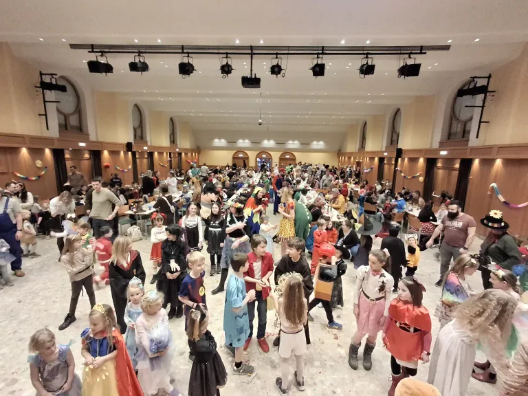 Eine große Menschenmenge von Kindern und Erwachsenen in Kostümen, die sich in einem Gebäude zu einer Veranstaltung versammelt haben, wahrscheinlich zu einer Kostümparty, wobei Erwachsene beaufsichtigen und einige Kinder tanzen oder spielen.