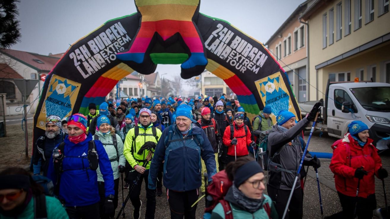 Eine Gruppe von Menschen, in Winterkleidung gekleidet, geht unter einem farbenfrohen Tor, mit einem Banner, das '24h Burgen Landextrem Tour' steht.