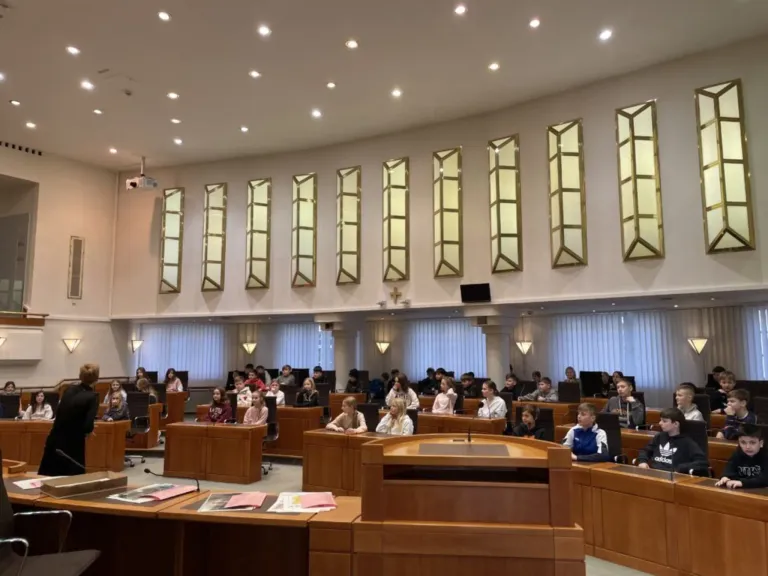 Ein Konferenzraum mit hölzernen Schreibtischen in Reihen, sitzenden Schülern, einem Podium vorne, einem Projektor und bogenförmigen Fenstern an der Wand.