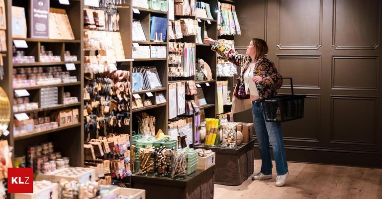Eine Frau steht in einem Boutique und zeigt auf ein Regal mit verschiedenen Artikeln. Sie hält einen Einkaufskorb und trägt Freizeitkleidung.