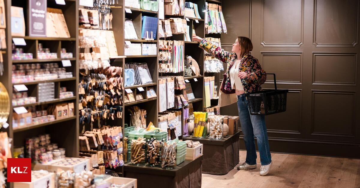 Eine Frau steht in einem Boutique und zeigt auf ein Regal mit verschiedenen Artikeln. Sie hält einen Einkaufskorb und trägt Freizeitkleidung.