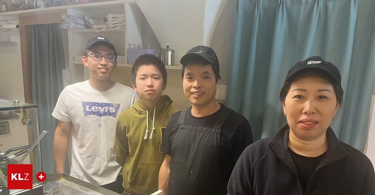 Vier Personen posieren für ein Foto in einer Küche. Ein Mann in einer Schürze lächelt, während andere Mützen und Hemden tragen. Dahinter befindet sich ein Regal mit Gegenständen.