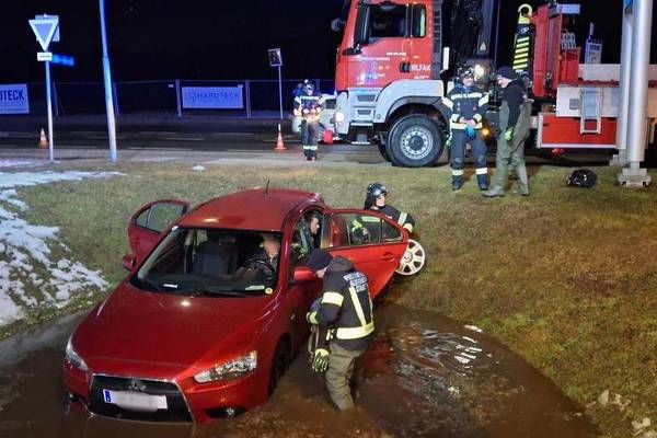 Ein rotes Auto ist in Wasser eingetaucht, mit Feuerwehrleuten und einem Feuerwehrwagen in der Nähe. Der Fahrer ist im Inneren, und einige Leute helfen.