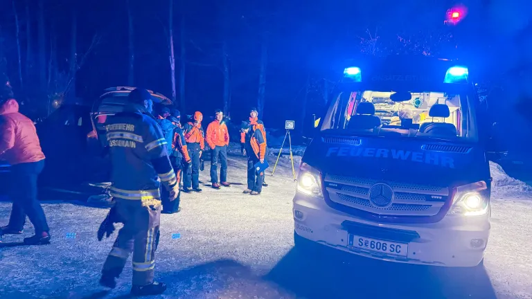 Eine nächtliche Szene mit Feuerwehrleuten in orangefarbenen Uniformen, die sich um einen Feuerwehrwagen mit eingeschalteten Lichtern versammelt haben. Die Feuerwehrleute scheinen sich in einem Waldgebiet zu befinden, mit Bäumen und einem blauen Himmel im Hintergrund.