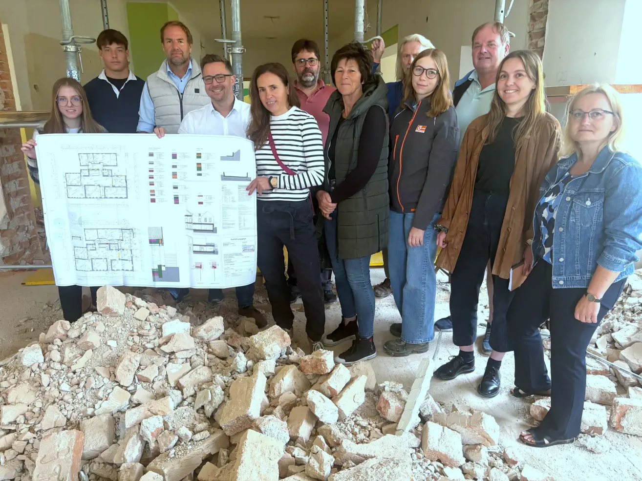 Eine Gruppe von Menschen steht an einer Baustelle mit einem Bauplan. Sie sind alle in Jacken, Hosen und Schuhen gekleidet. Einige tragen Brillen. Der Boden ist mit Schutt gefüllt.