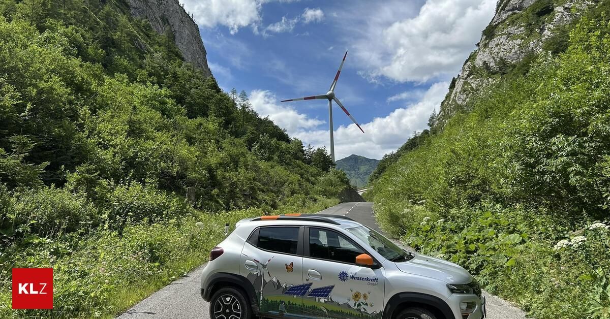 Ein silberner Wagen ist auf einer Bergstraße geparkt, mit einem Windrad und üppiger Grünfläche im Hintergrund.