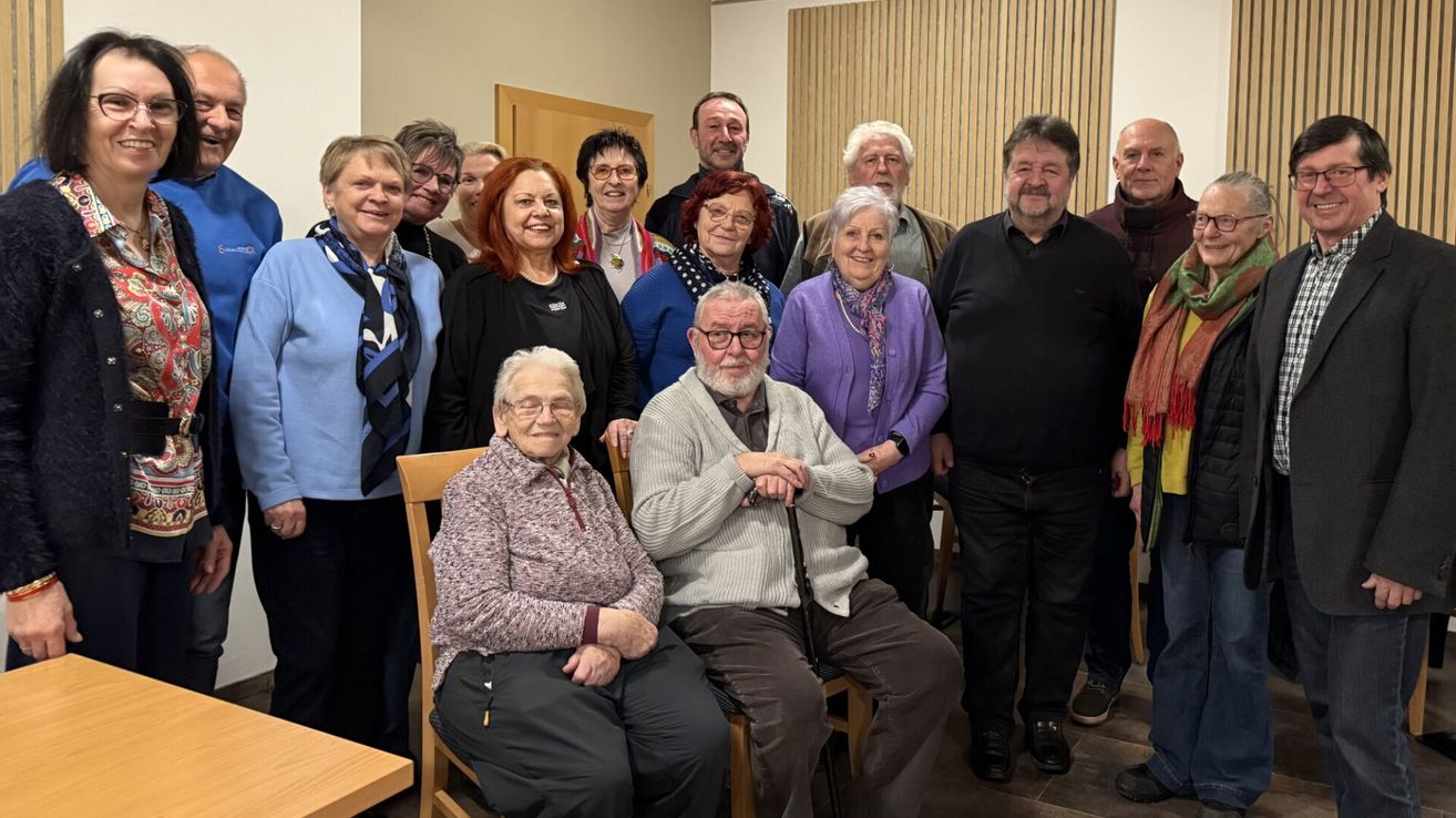 Eine Gruppe von Menschen, meist ältere Personen, posiert für ein Foto. Sie alle lächeln und scheinen glücklich zu sein. Der Raum hat Holzwände und einen Holzboden.