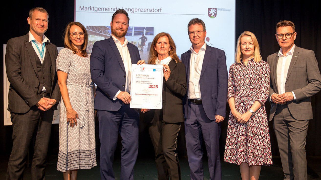 Fünf Personen stehen auf einer Bühne und posieren für ein Foto. Eine Frau hält ein Zertifikat mit der Aufschrift 'Zertifikat 2025'. Hinter ihnen zeigt eine Leinwand den Text 'Marktgemeinde Langenzersdorf'.