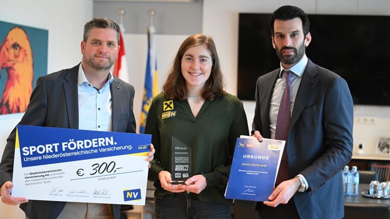 Drei Personen stehen vor einer Wand mit Fahnen und halten Auszeichnungen. Eine Frau hält eine Trophäe, ein Mann links von ihr hält ein blaues Zertifikat und ein Mann rechts von ihr hält ein blaues Zertifikat. Die Wand hinter ihnen hat Fahnen und einen Fernsehbildschirm.