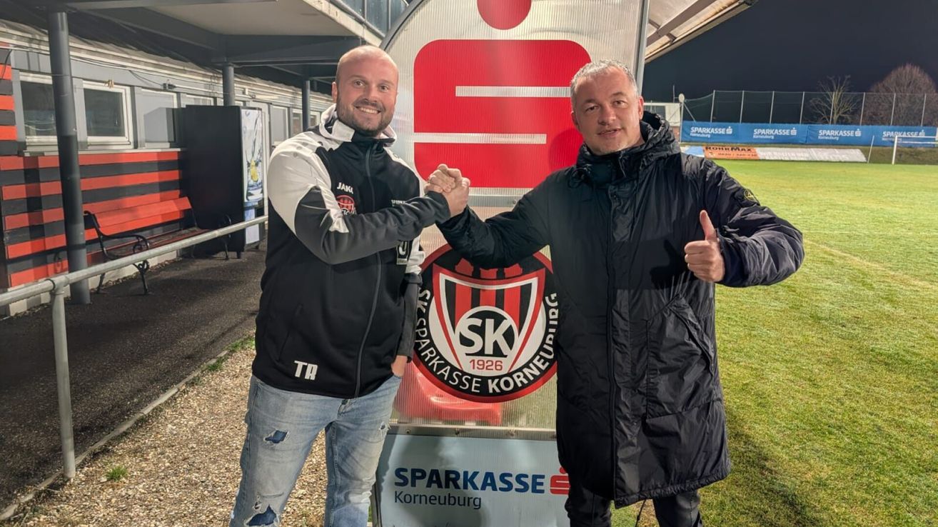 Zwei Männer schütteln sich die Hände vor einem Schild von Sparkasse Korneburg. Einer trägt eine schwarz-weiße Jacke, der andere eine schwarze Jacke. Beide lächeln.
