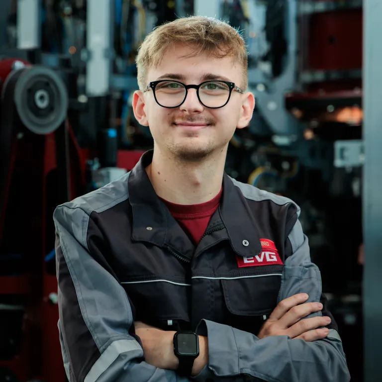 Ein junger Mann mit Brille steht lächelnd in einer Werkstatt, trägt eine graue und schwarze Jacke mit einem roten Logo auf der linken Brust. Hinter ihm sind Industriemaschinen sichtbar.