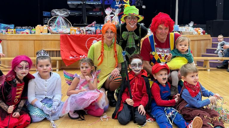 Eine Gruppe von Kindern und Erwachsenen in Kostümen posiert für ein Foto. Dahinter ist ein Hintergrund mit einem Herz und Spielzeug.