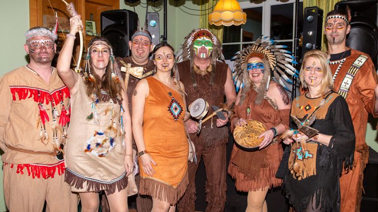 Eine Gruppe von Menschen in indianischen Kostümen, die lächelnd für ein Foto posieren. Einige tragen Gesichtsbemalung und halten Trommeln. Ein Lautsprecher und eine Lampe sind im Hintergrund zu sehen.