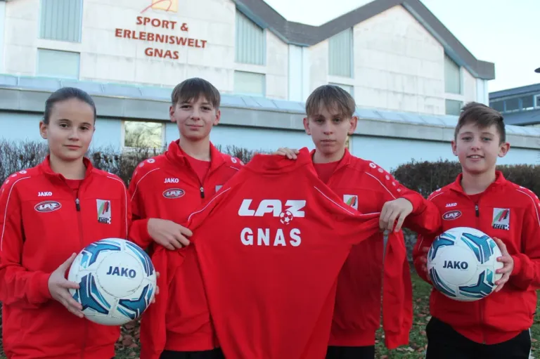 Vier Jungen in roten Sporttrikots halten ein rotes Shirt mit dem Wort GNAS hoch. Sie stehen vor einem Gebäude mit einem Schild, auf dem Sport & Erlebniswelt GNAS steht.