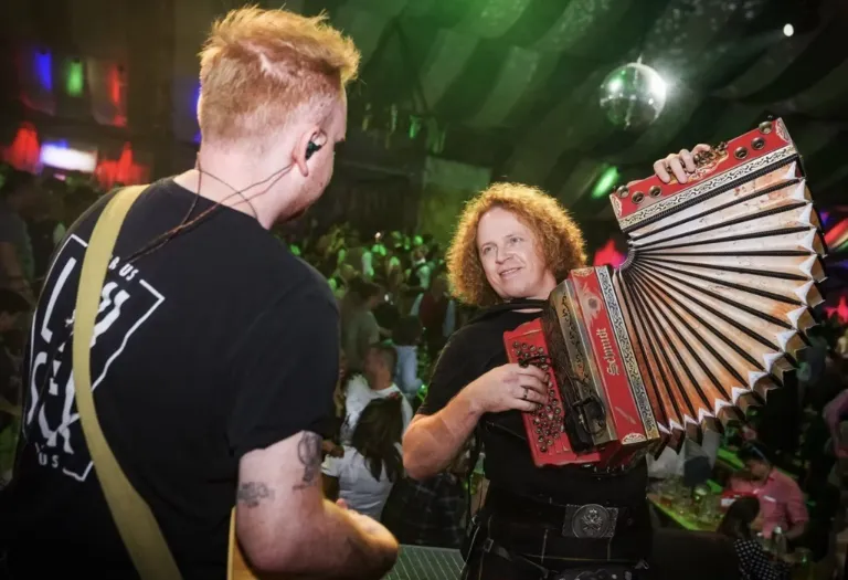 Eine Frau spielt ein rotes Akkordeon in einer Bar, mit einem Mann mit Kopfhörern hinter ihr und einer Menschenmenge im Hintergrund.