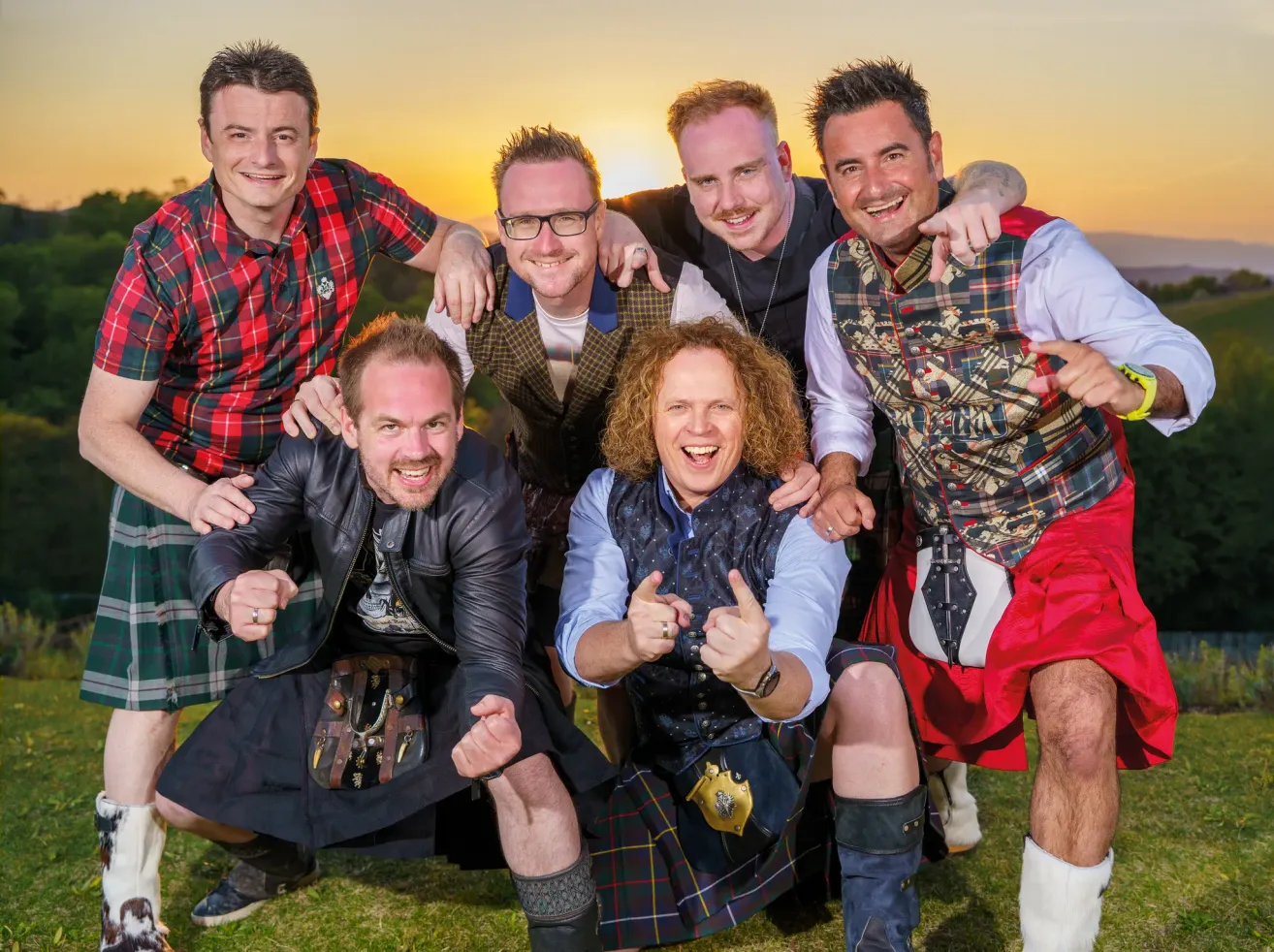 Eine Gruppe von Männern in Kilts und Westen posiert für ein Foto auf einem Rasenfeld, wobei zwei Männer in der Mitte Daumen hoch halten.