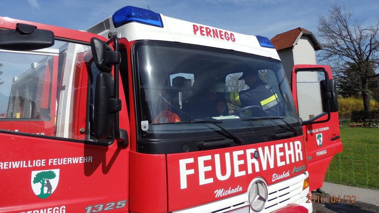 Ein roter Feuerwehrwagen mit 'PERNEGG' auf der Oberseite und 'FEUERWEHR' an der Seite. Er hat eine blaue Licht auf der Oberseite und die Nummer 1325. Zwei Personen sind im Inneren, eine fährt und eine sitzt.