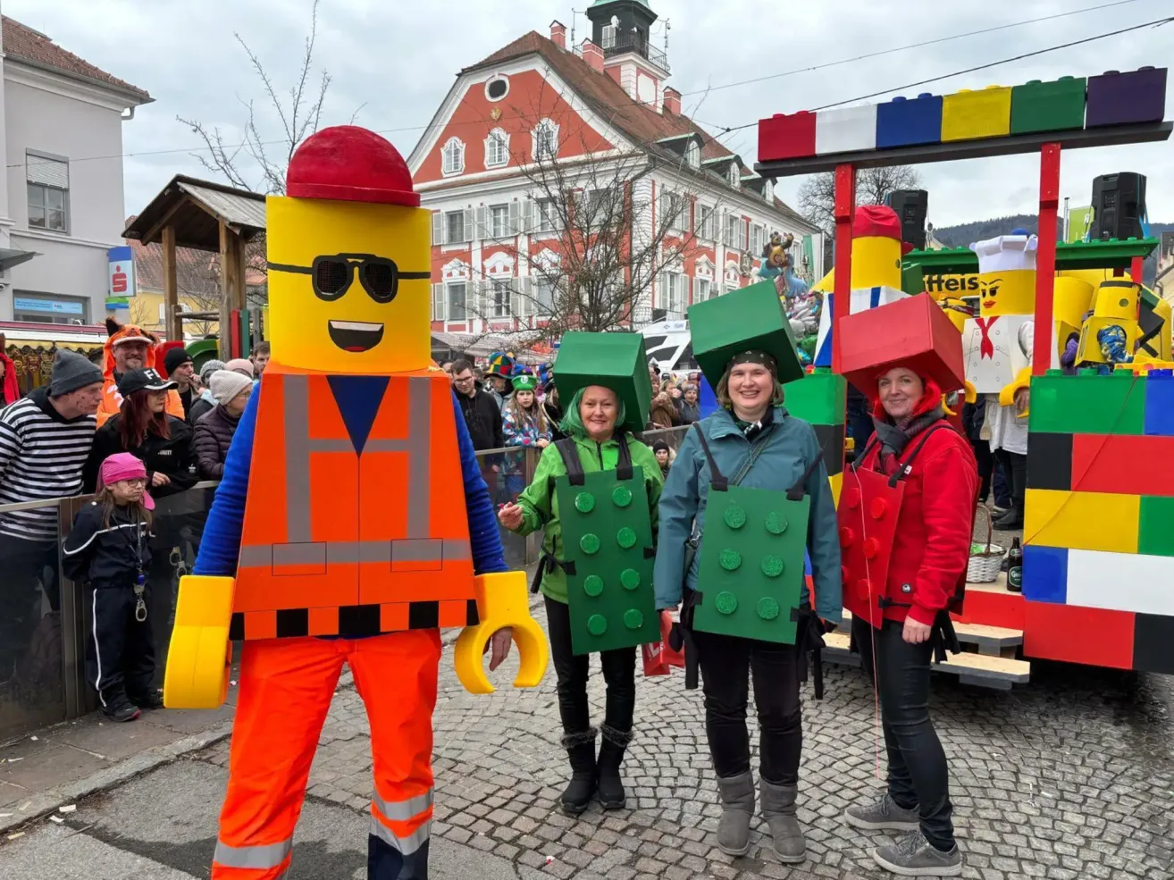 Vier Menschen in Lego-Kostümen stehen vor einer bunten Struktur. Dahinter versammelt sich eine Menschenmenge, und ein Gebäude mit Turm ist sichtbar.