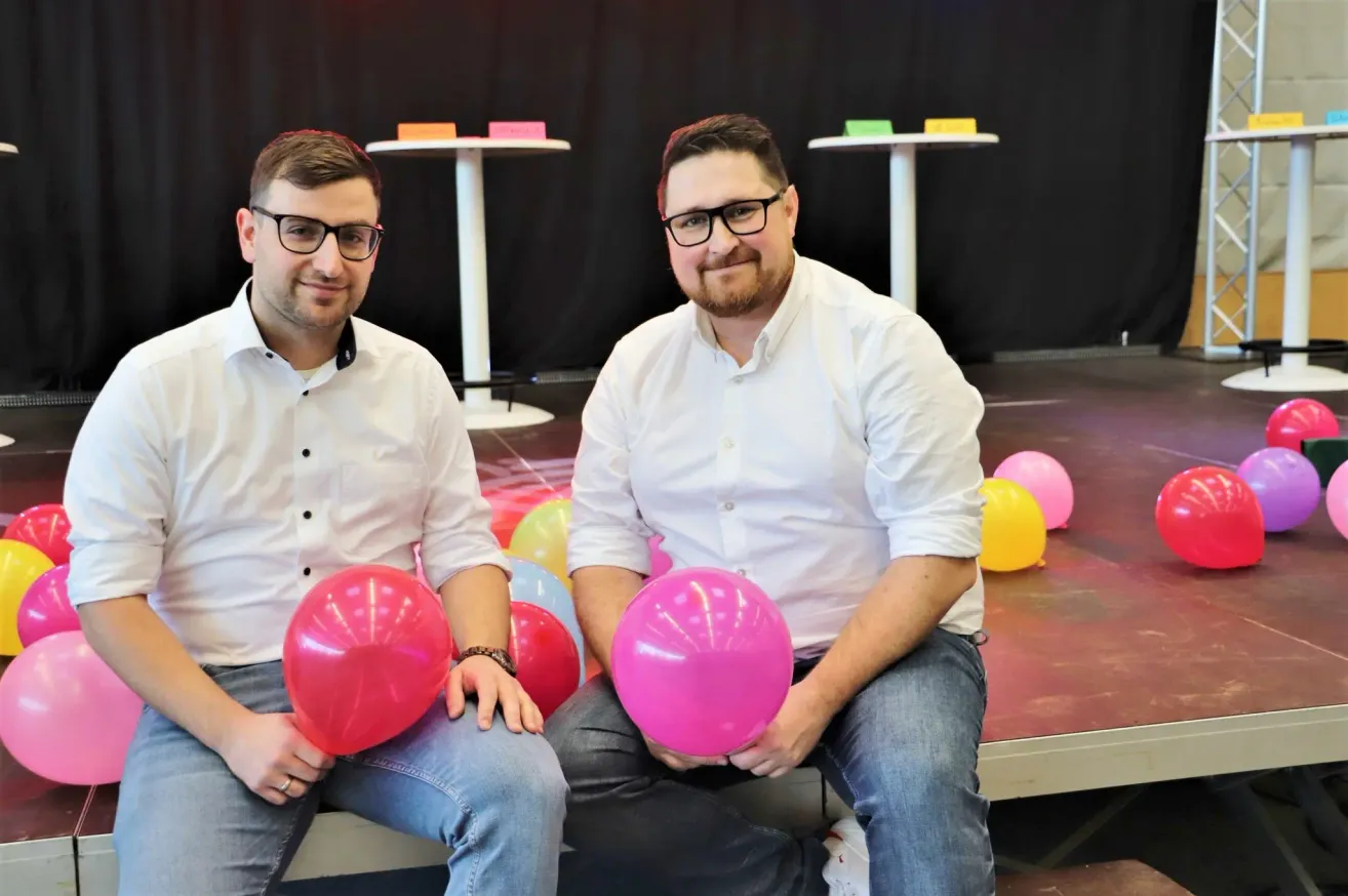 Zwei Männer sitzen auf einer Bank und halten Luftballons. Einer trägt eine Brille und der andere einen Bart. Sie sind von bunten Luftballons und Tischen mit bunten Haftnotizen umgeben.