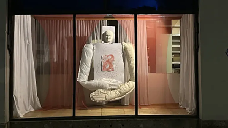 Eine weiße Statuenfigur mit einem roten Herz auf weißem Hintergrund ist in einer Glasvitrine mit roten Vorhängen an den Seiten ausgestellt.