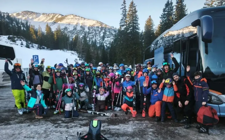 Eine Gruppe von Skifahrern und Snowboardern, sowohl Erwachsene als auch Kinder, posiert für ein Foto vor einem verschneiten Berg. Sie tragen farbenfrohe Winterausrüstung und Helme, mit Skiern und Stöcken um sie herum. Ein Bus ist im Hintergrund zu sehen.