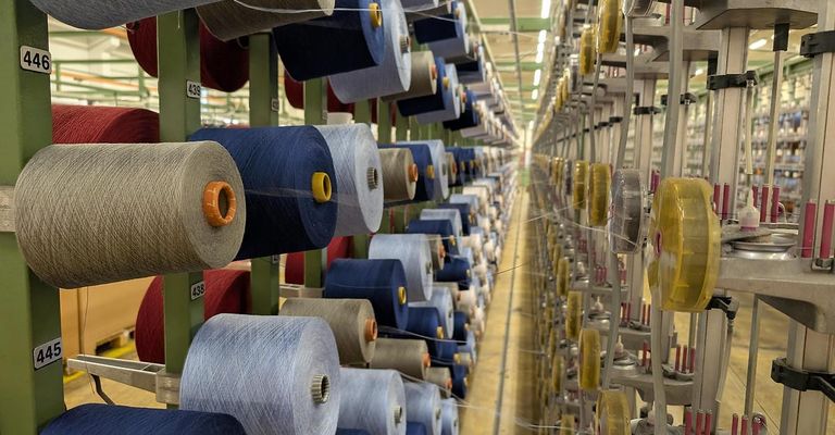 Ein Fabrikboden gefüllt mit zahlreichen Spulen von Garn in verschiedenen Farben, ordentlich auf Metallregalen unter heller Beleuchtung organisiert. Die Spulen sind mit Nummern gekennzeichnet und haben gelbe und orangefarbene Kappen.