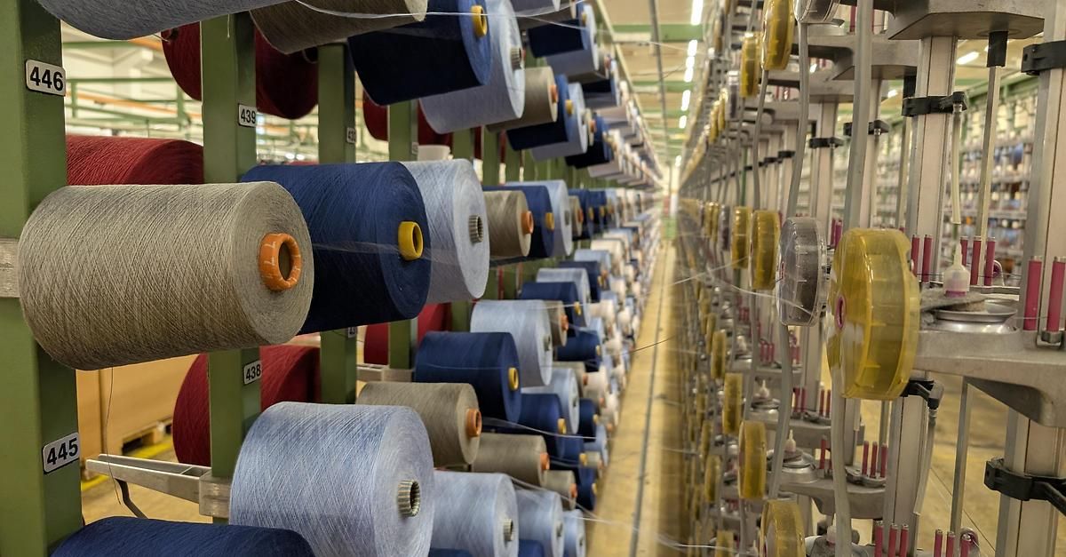 Ein Fabrikboden gefüllt mit zahlreichen Spulen von Garn in verschiedenen Farben, ordentlich auf Metallregalen unter heller Beleuchtung organisiert. Die Spulen sind mit Nummern gekennzeichnet und haben gelbe und orangefarbene Kappen.